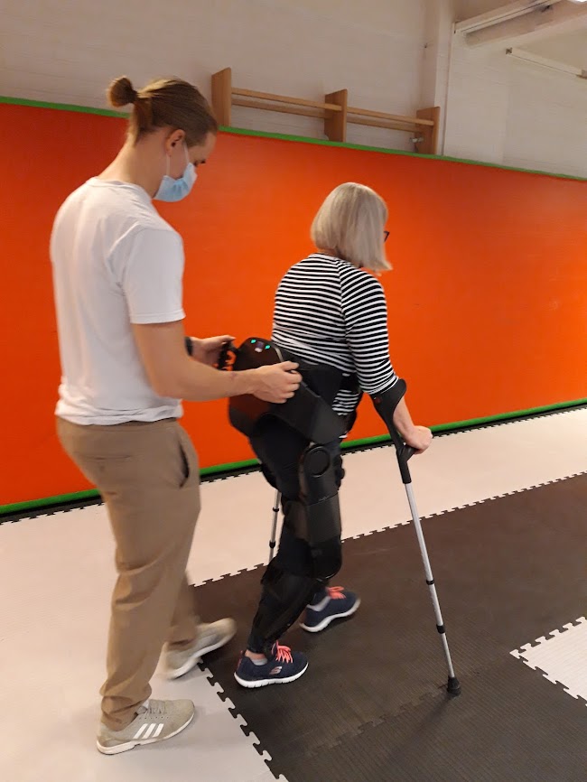 Kuvassa naispotilas käyttää Indego eksoskeletonia ja hänen takanaan on ohjaaja.