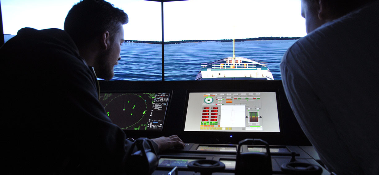 SAMK Simulator, two people drive on a simulator ship. In the front is the cockpit, in the background is the bow of the ship.