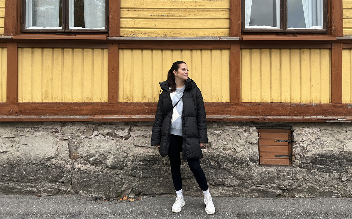 Ellen Lyyski seisoo vanhan puutalon vieressä ja katsoo sivulle.