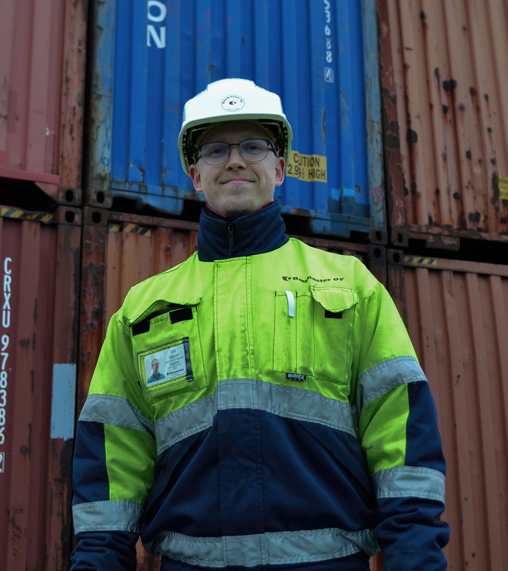 Olli Viljanen seisoo konttien edessä keltaisessa turvatakissa ja valkoisessa kypärässä.