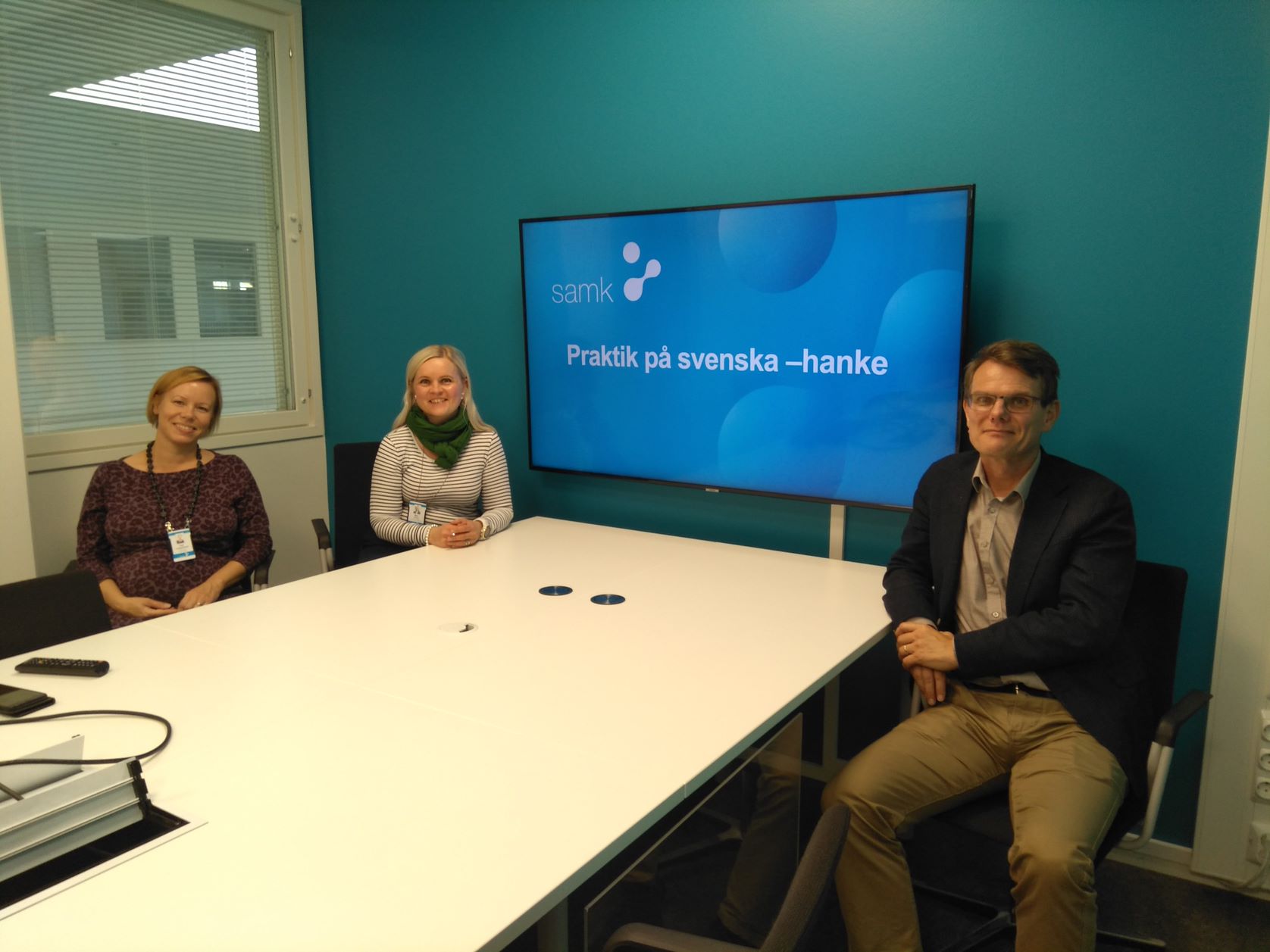 Hankkeen ihmiset istuvat yhdessä pöydän ääressä. Taustalla televisiossa näkyy hankkeen nimi Praktik på svenska.