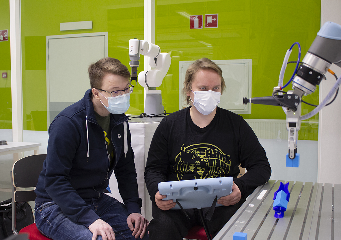 Kuvassa kaksi miestä käyttävät yhteistyörobottia pendantin avulla.