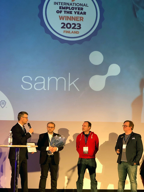 Neljä miestä seisoo lavalla palkisemistilaisuudessa. Four men stand on stage at the awards ceremony.
