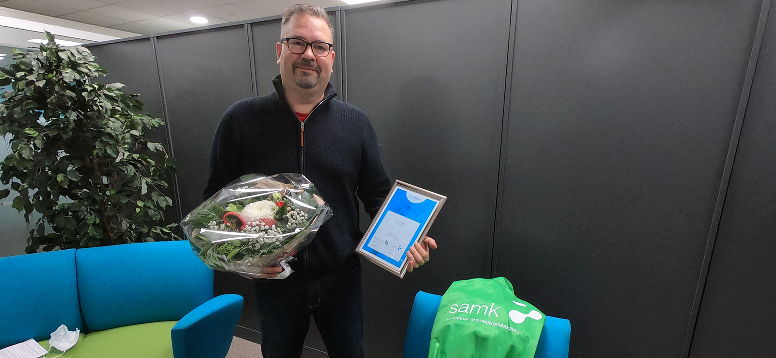 Petri Martikkala stands in front of a gray border, holding a certificate of honor and a bouquet of flowers.