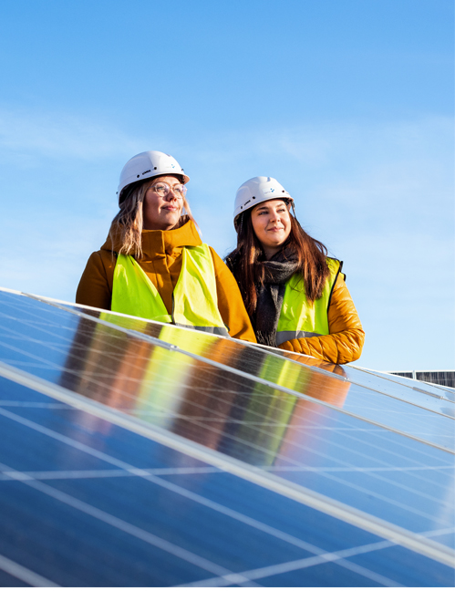 Two students stand behind the solar panels, wearing safety clothing.