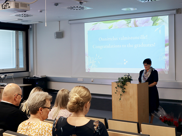 Naishenkilö puhuu edessä puhujapöntön takana valmistujaistilaisuuteen osallistuville.