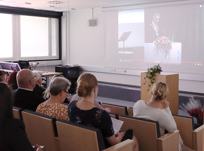 Valmistujaistilaisuuteen osallistuvat katsovat screeniltä tulevaa puhetta.