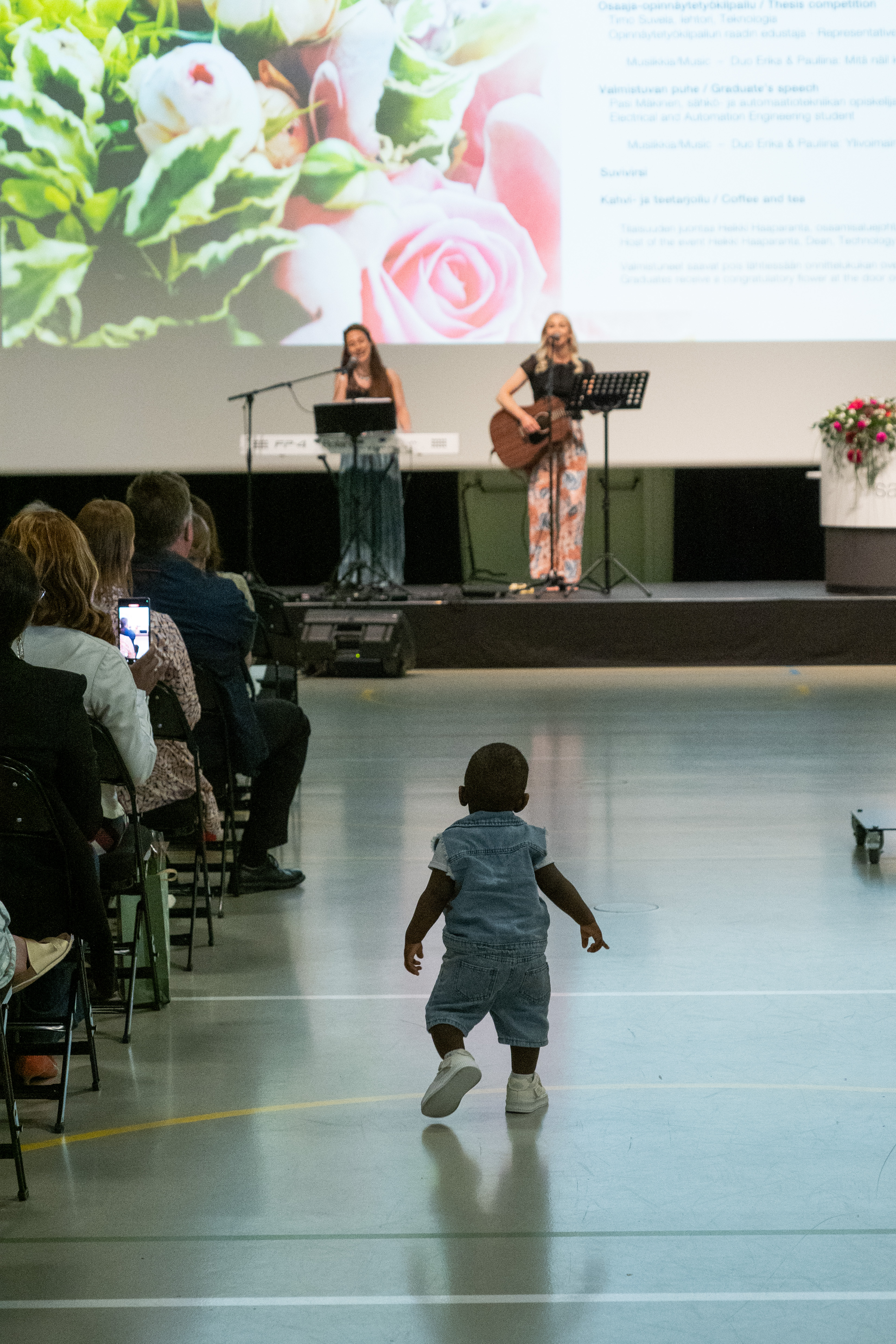 A little child walks down the aisle and in the background two women perform on stage with a guitar and a keyboard.