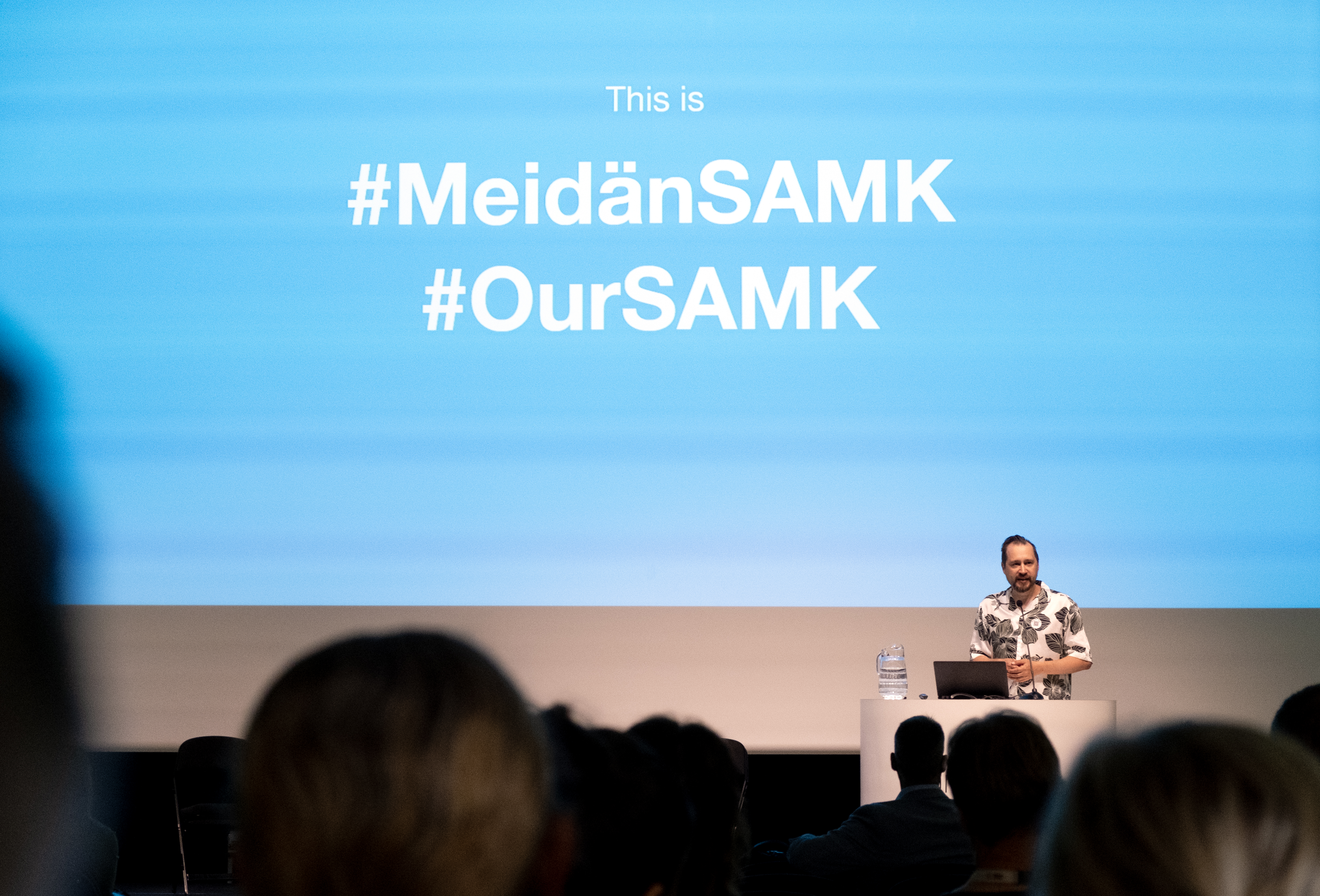 A man stands in a lectern in front of a large screen.