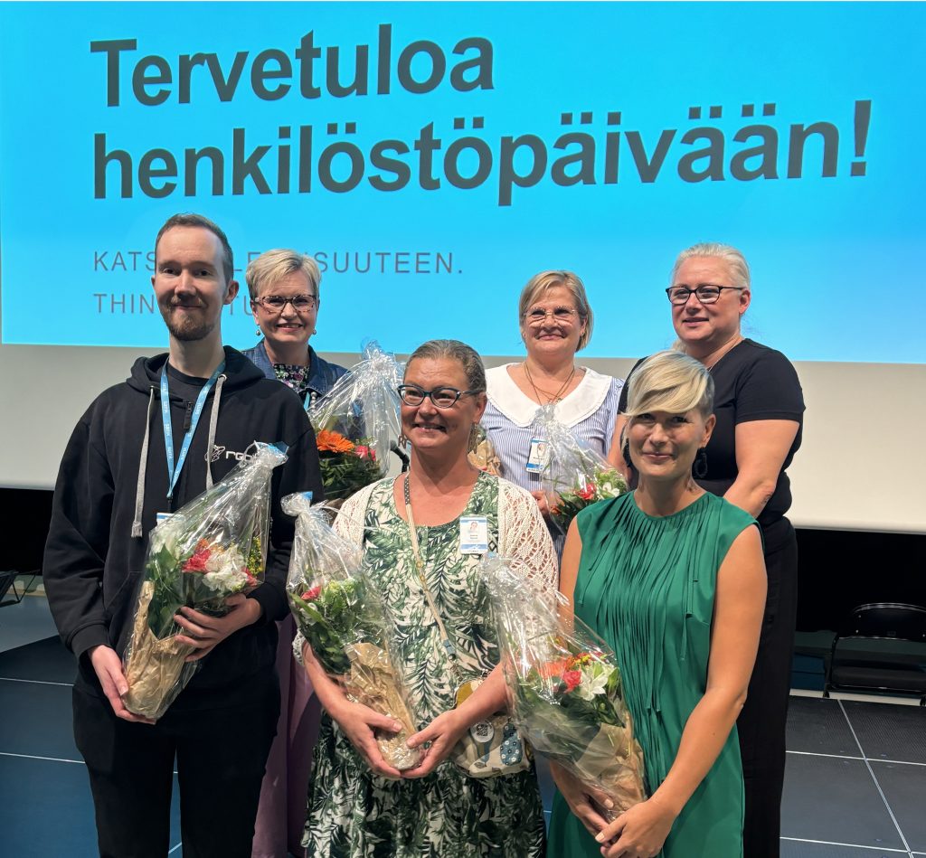 A man and five women are standing in front of a large screen with bunches of flowers in their hands.