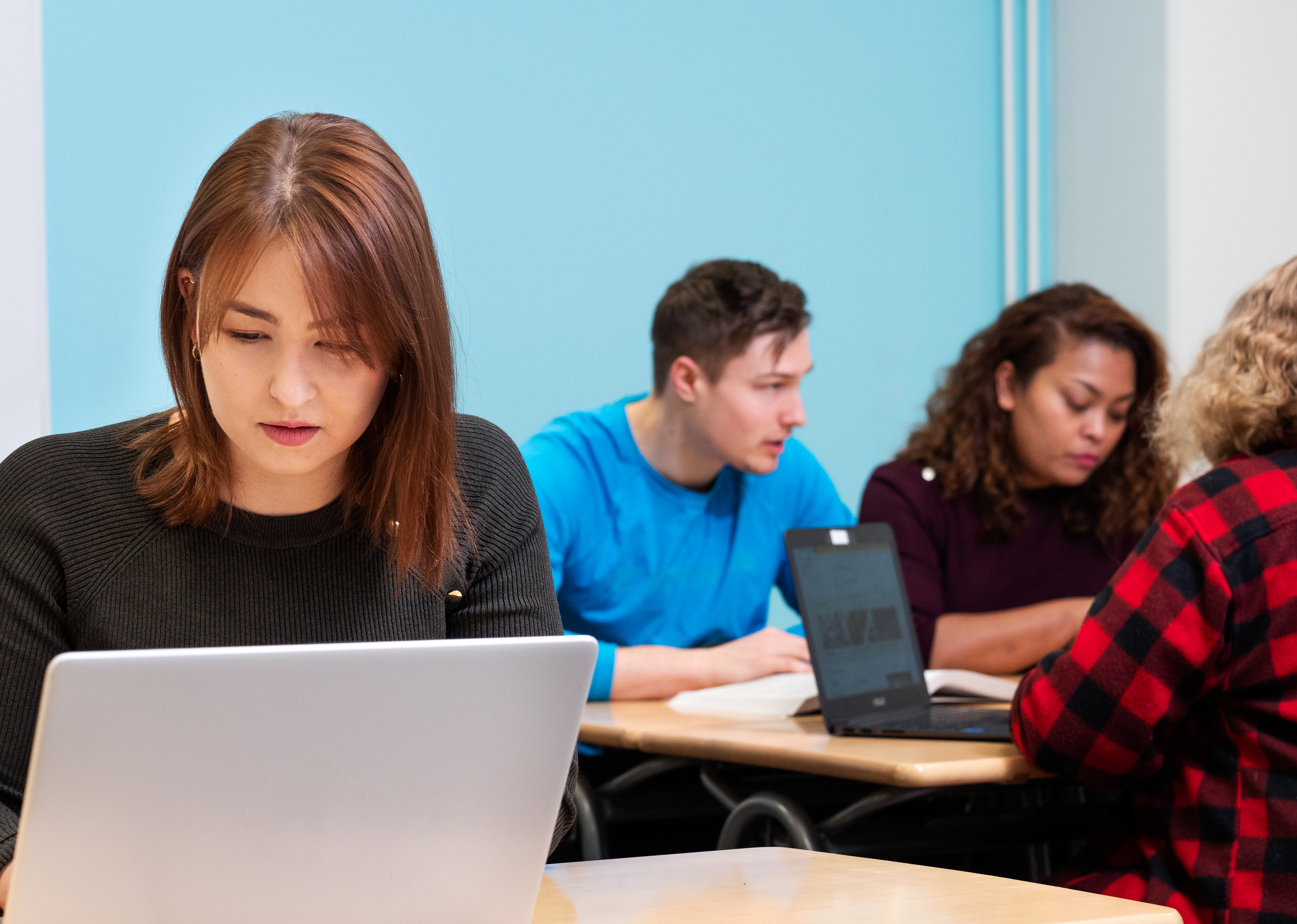 A female student is sitting at a laptop with a group of students in the background.