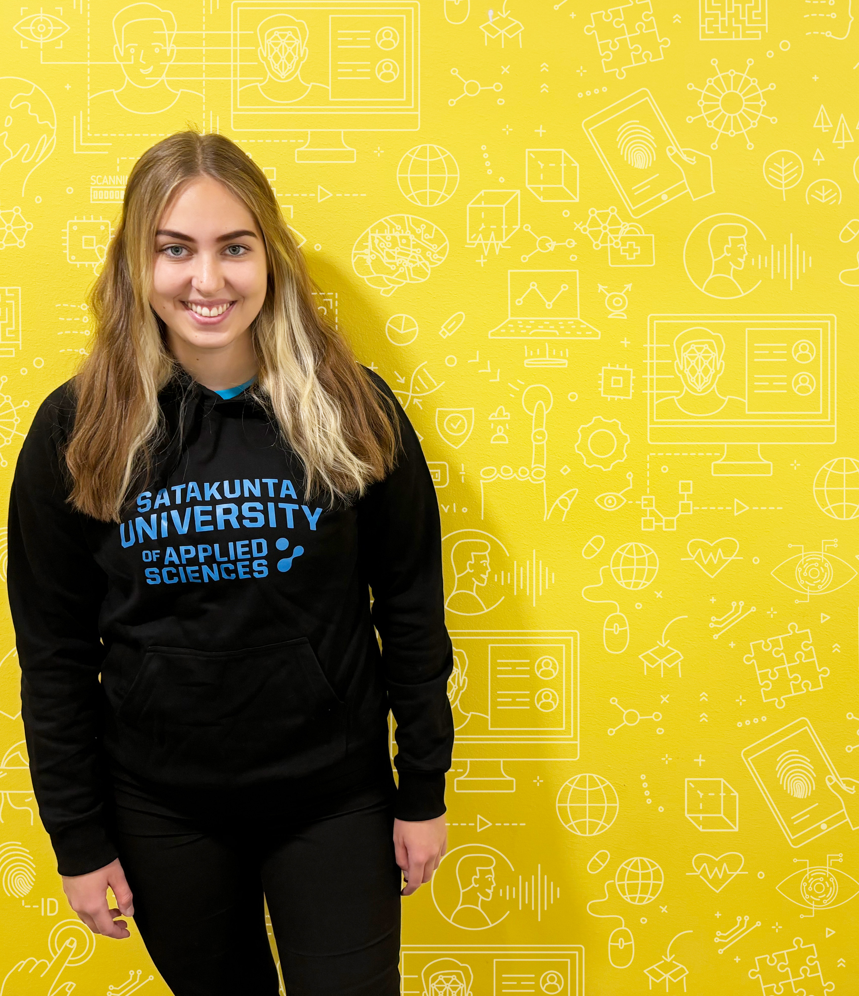 A student standing in front of a yellow wall wearing a SAMK hoodie.