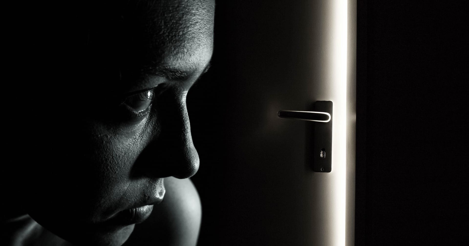 Nainen katsoo mustavalkoisessa kuvassa pelokkaasti ovelle, jonka raosta näkyy valoa.