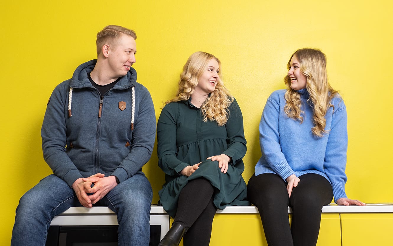 Mies ja kaksi naista istuu keltaisen seinän edessä ja juttelee keskenään.