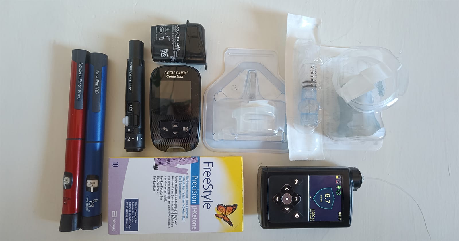 Valokuva tyypin 1 diabeteksen hoidossa käytettävistä välineistä tasaisella alustalla. Kuvassa on muun muassa kaksi monikäyttöistä NovoPen-insuliinikynää (pitkävaikutteiselle ja ateriainsuliinille), Accu-Chek-verensokerimittari, Freestyle-ketoainemittausliuskoja, insuliinipumpun tarvikkeita sekä nykyaikainen MiniMed 780G-älyinsuliinipumppu, jonka näytössä näkyy verensokerilukema 6,7 mmol/l.