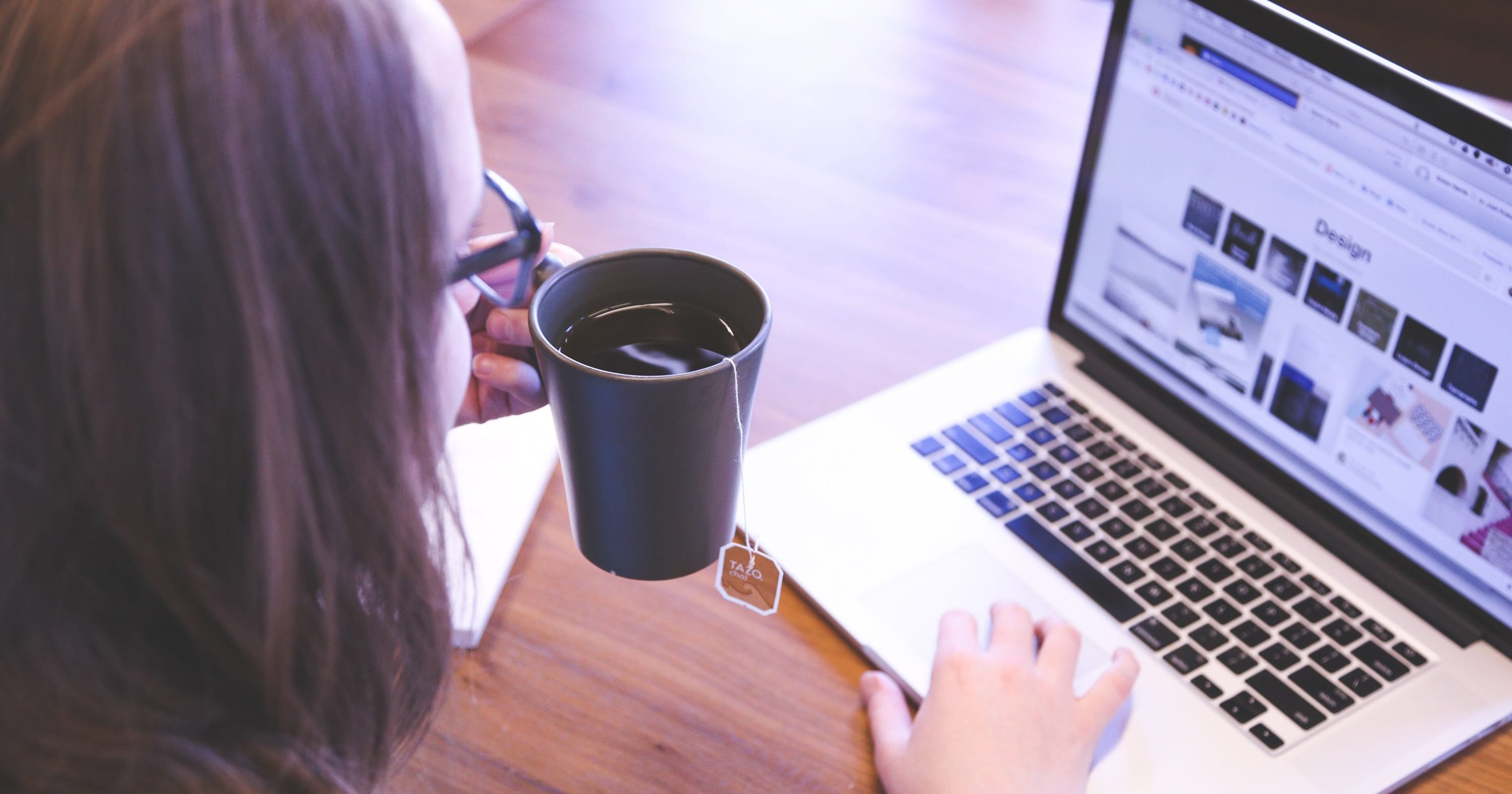 Nainen on teemuki kädessä kannettavan tietokoneen ääressä. The woman is sitting at a laptop with a mug in her hand.