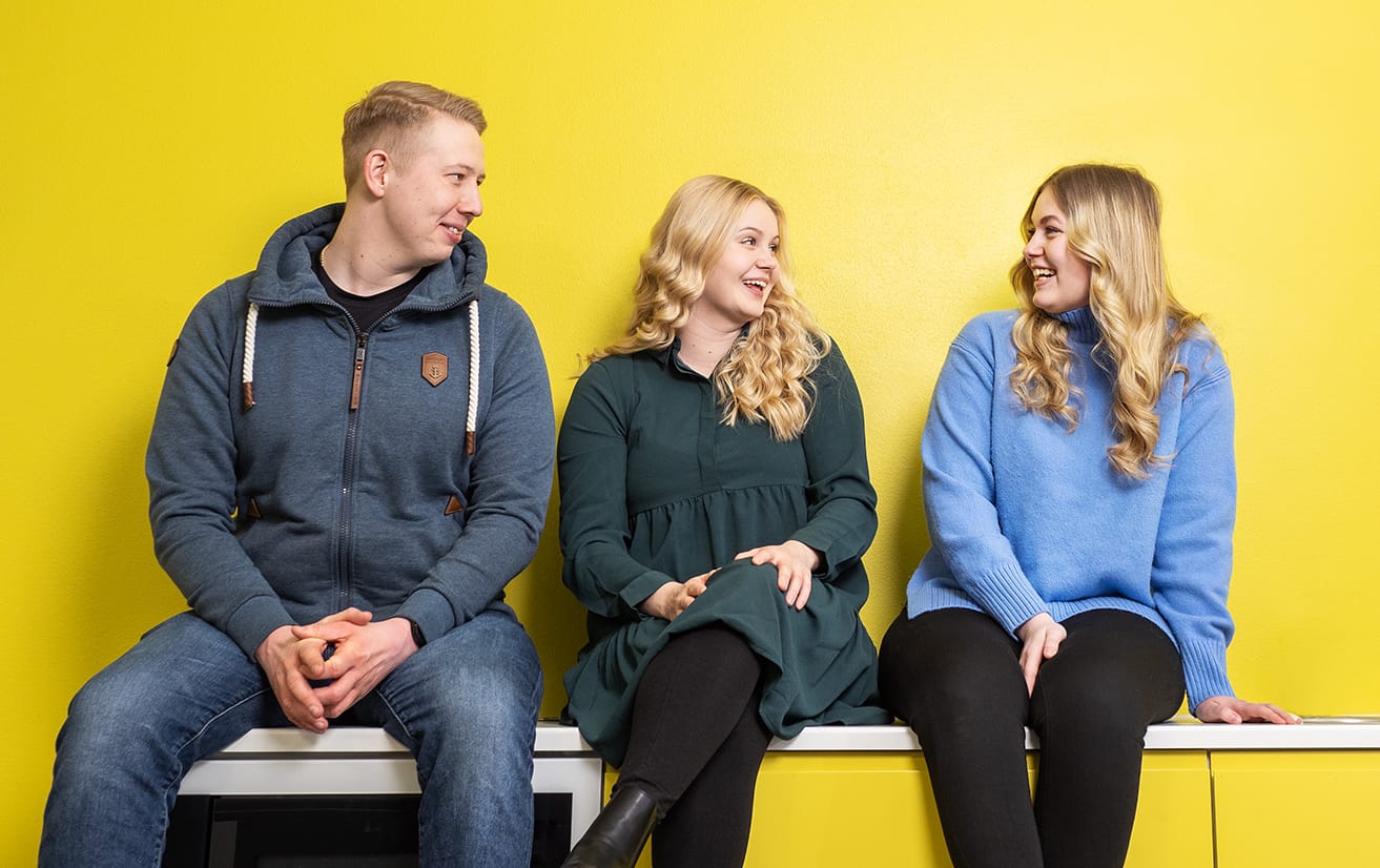 Kolme opiskelijaa istuu keltaisen seinän edessä.