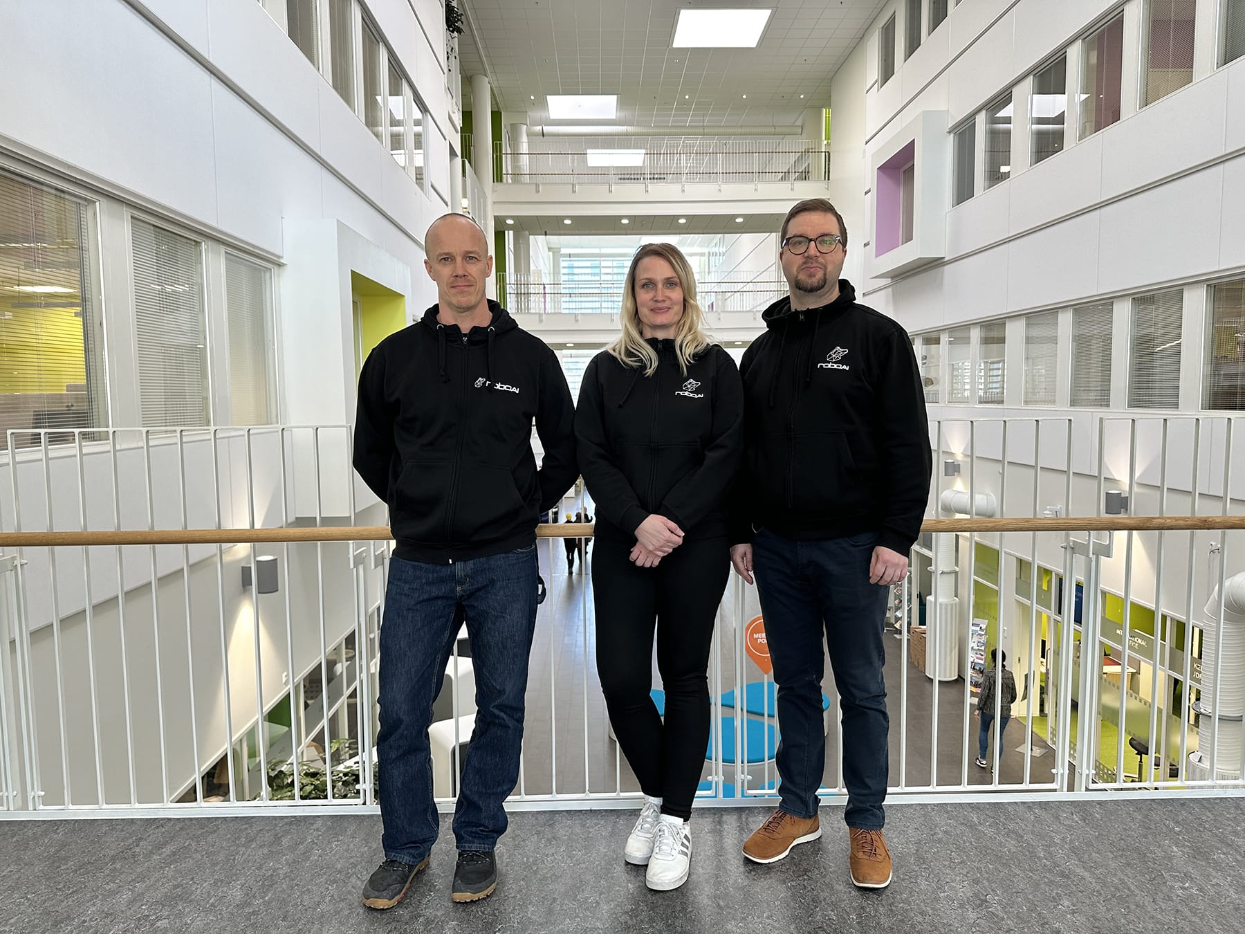 Three people are standing on an indoor mezzanine wearing matching work hoodies.
